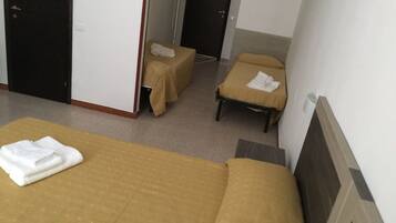 A hotel room with two single beds, each with a brown bedspread and a white towel. There are two small tables with brown tablecloths, one in front of each bed. The room has white walls and a tiled floor.