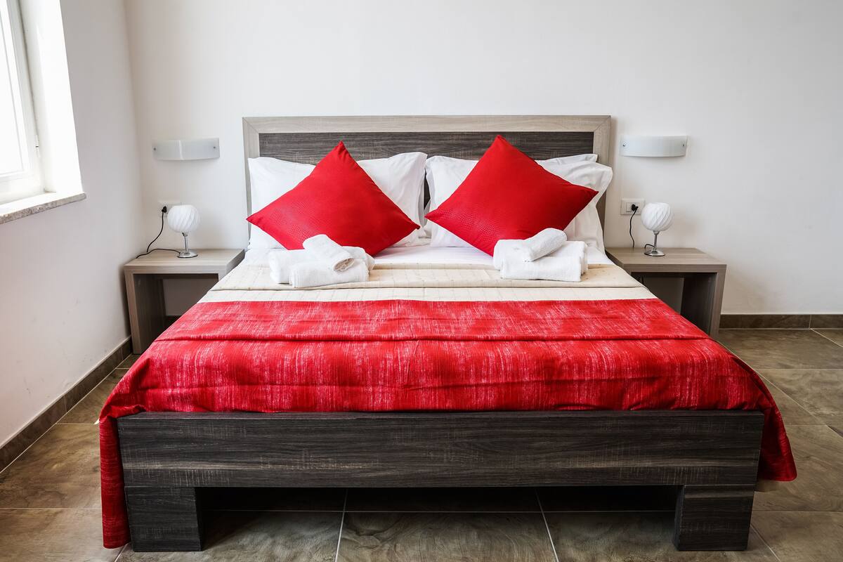 A neatly made bed with a red and white bedspread, two red pillows, and white towels on a dark wooden bed frame.