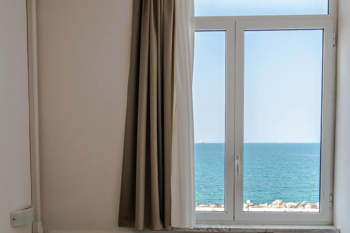 A bedroom with a bed, a nightstand with a lamp, a window with curtains, and a view of the sea.