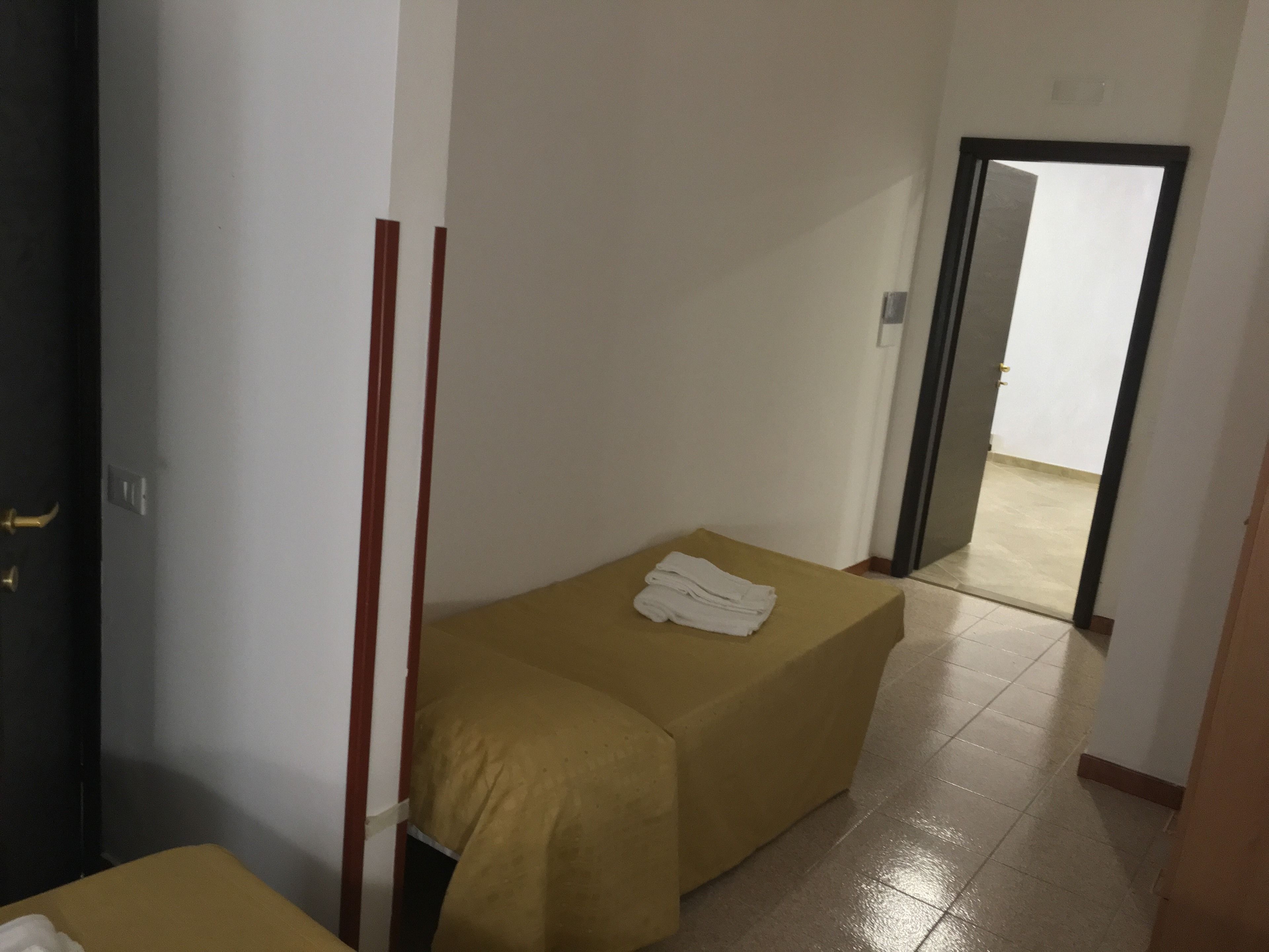 A small, simple room with a bed and a bench. There is a door leading to another room.