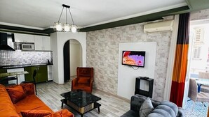 Ein modernes Wohnzimmer mit Sofa, Sessel und Couchtisch. Es gibt eine Küchenzeile mit Mikrowelle und einem Esstisch. Ein Fernseher ist an der Wand befestigt, und eine Klimaanlage ist zu sehen.