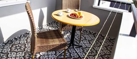 A small outdoor seating area with a wicker chair and a round table, featuring a plate of food and a cup.