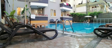 Una piscina interior con sillones de descanso, suelo de baldosas y un edificio con balcones al fondo.