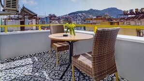 Ein Balkon mit gemustertem Boden, Korbstühlen, einem runden Tisch mit Blumenvase und Blick auf eine Stadtlandschaft mit Gebäuden und Hügeln im Hintergrund.