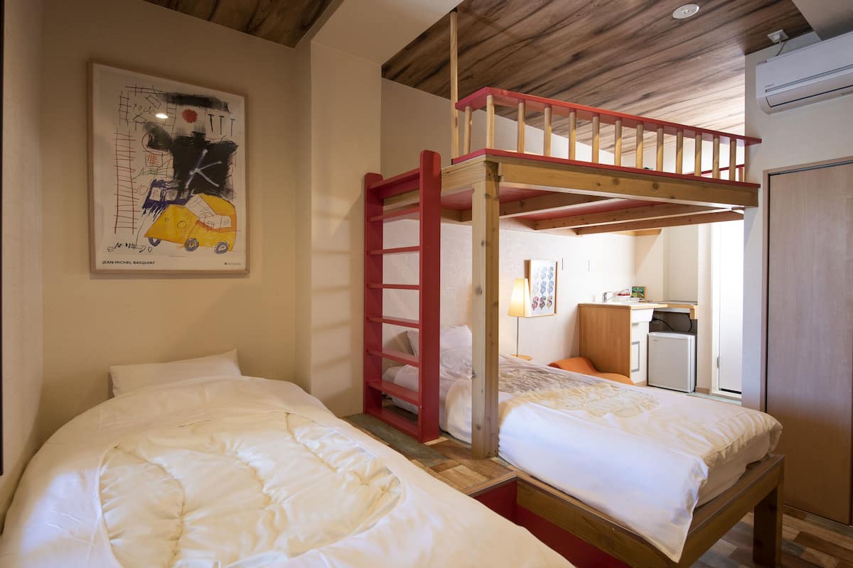 A room with two single beds, a bunk bed, a wooden ceiling, and a kitchen area in the background.