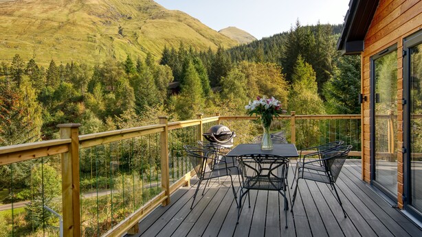 En træterrasse med et bord og stole, med udsigt over et bjergrigt landskab med træer.