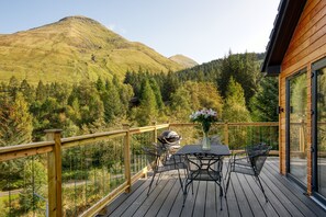 Terraza de madera con mesa y sillas, con vista a un paisaje montañoso y árboles.