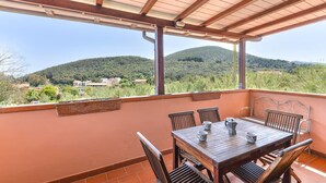 Une terrasse extérieure couverte, avec une table et des chaises en bois, une vue sur un paysage montagneux et un mur de couleur terre cuite.