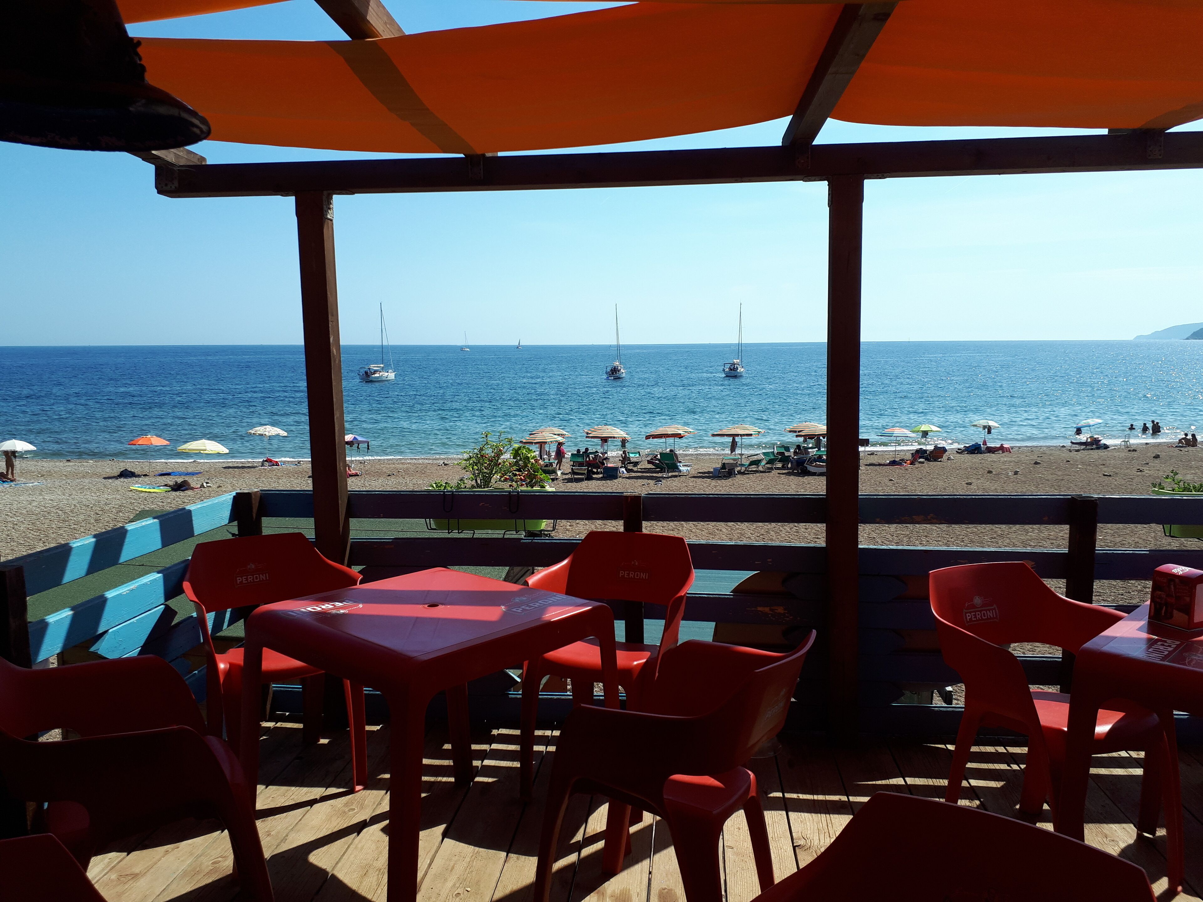 A beachside restaurant with outdoor seating, a view of the sea, and boats in the distance.