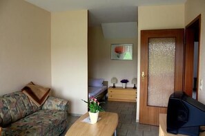 Een knus woongedeelte met een bank, een klein tafeltje met een plant, een bed en een deur met een reliëfpaneel.