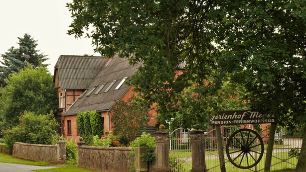 A traditional brick building with a sloped roof, a stone wall, and a gate with a large wheel. - Ferienhof Meyer (Nahrendorf)