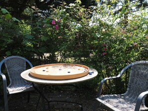 Een ronde tafel met een spel, twee rieten stoelen en een tuin met verschillende planten en bloemen.