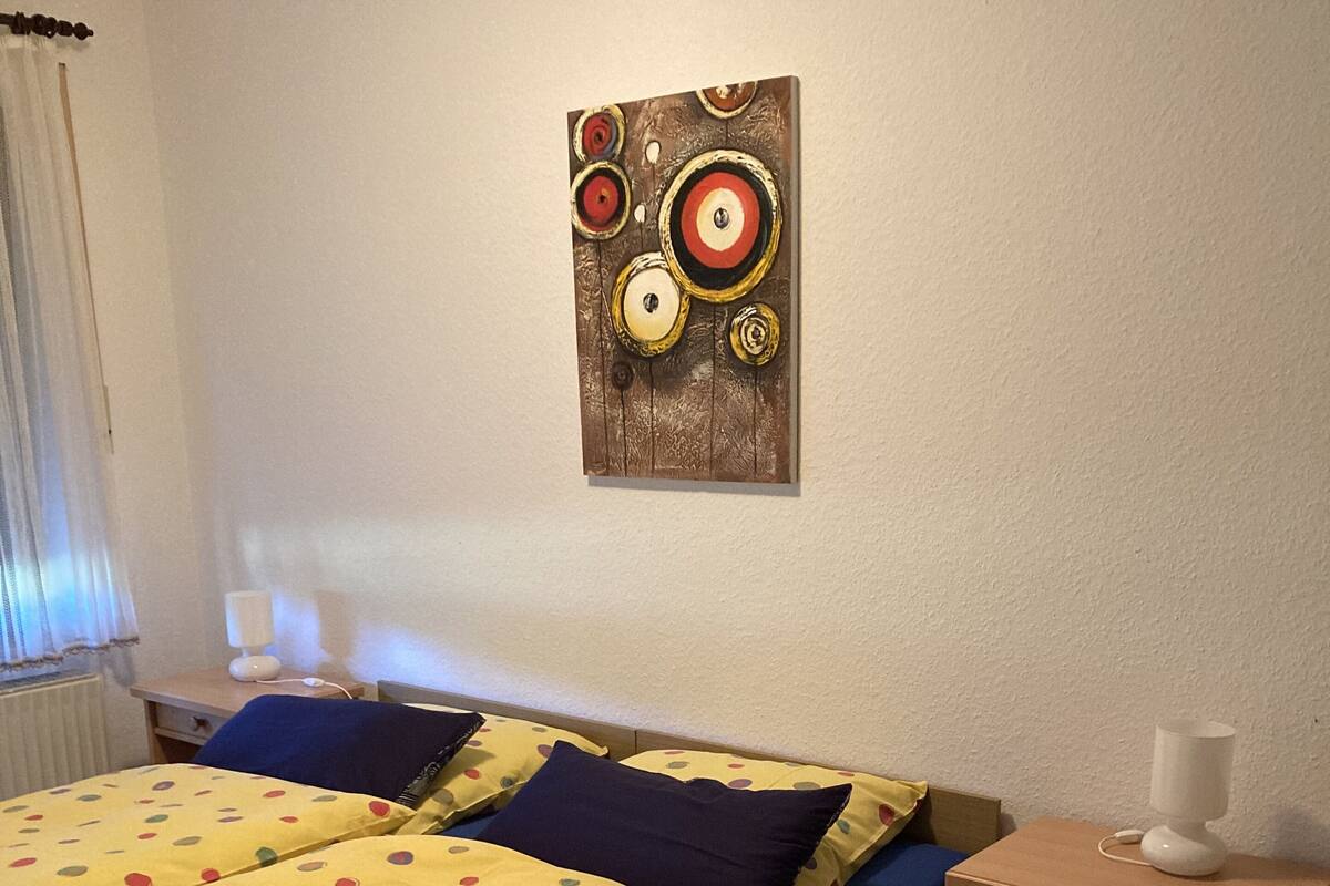 Una habitación con cama, mesita de noche, lámpara y una obra de arte en la pared.