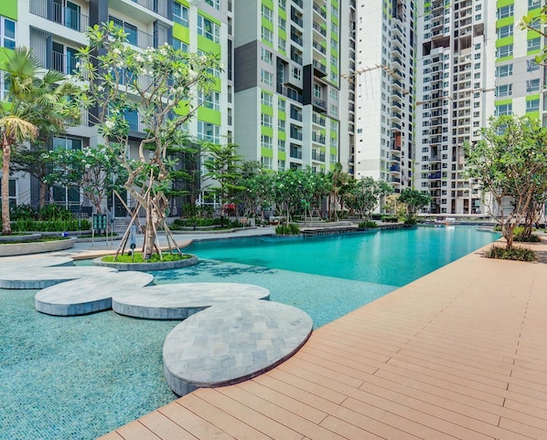 A modern hotel with a swimming pool, surrounded by greenery and a multi-story building in the background.