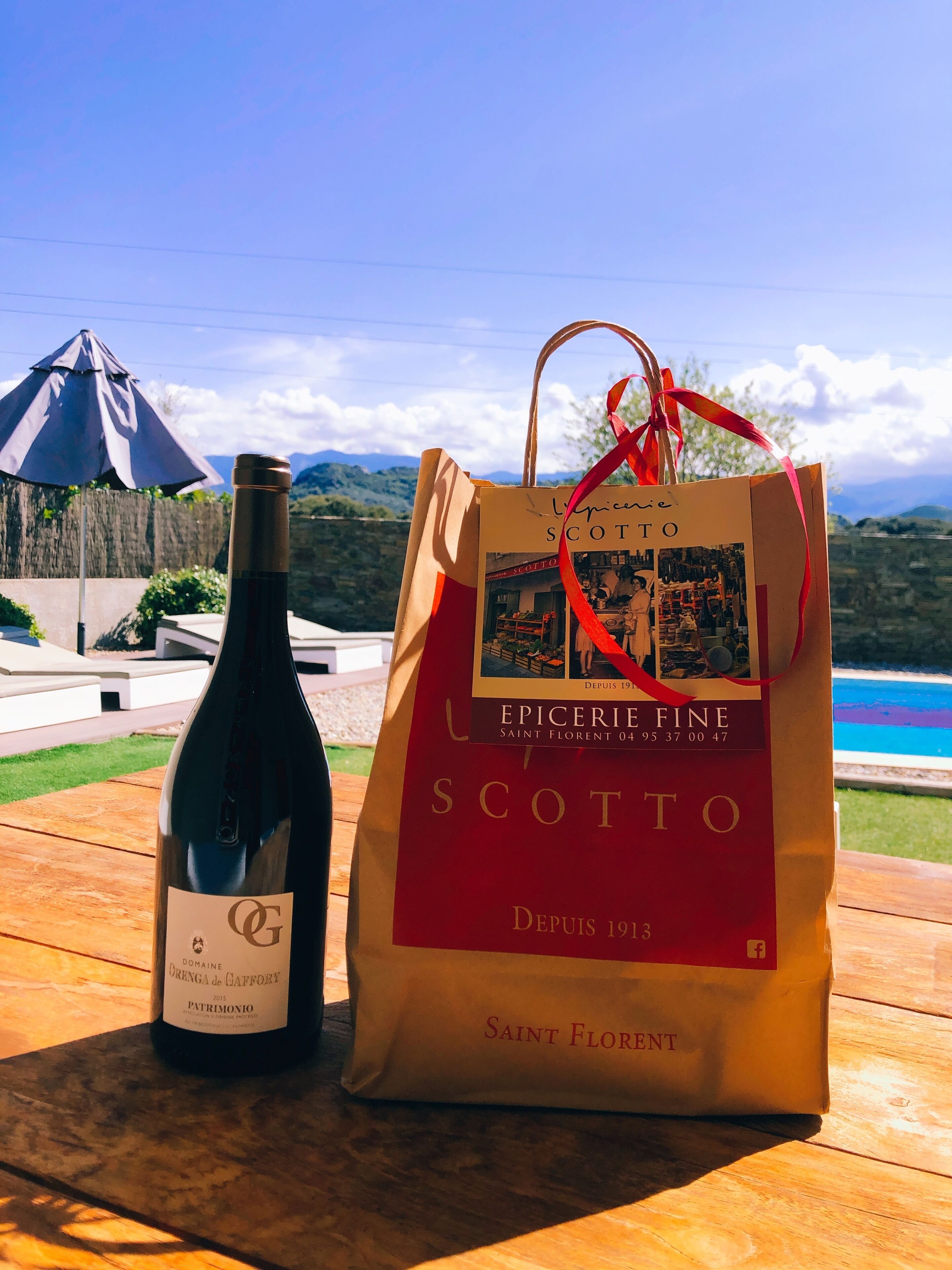 A bottle of wine and a gift bag on a wooden table with a scenic view of mountains and a pool in the background.