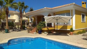Ett område vid poolen med en klarblå pool, en gul byggnad med en täckt uteplats och palmer.