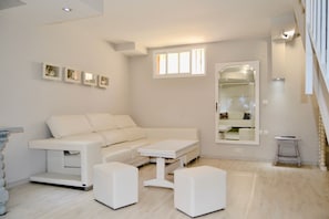 A modern living room with a white sofa, a coffee table, and two ottomans. There are framed pictures on the wall and a large mirror.