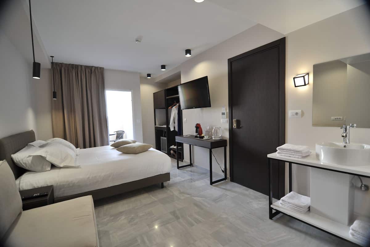 A modern hotel room with a large bed, a bedside table, a flat-screen TV, a dark wooden door, and a bathroom with a white sink and towel rack.