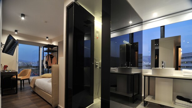 Habitación de hotel moderna con cama, escritorio, silla, televisor, puerta de vidrio, baño con lavamanos y espejo, y vista a la ciudad.