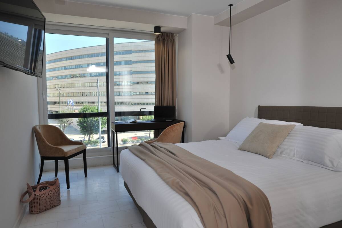 A hotel room with a bed, a desk, a chair, and a large window offering a view of a modern building.