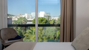 Habitación de hotel con un ventanal grande, una cama, una silla y vistas a edificios y zonas verdes.