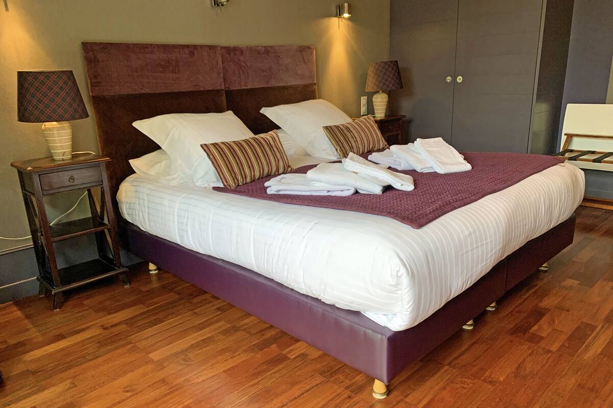 A neatly made bed with white linens and a purple bedspread, flanked by two nightstands with lamps, in a room with wooden flooring.