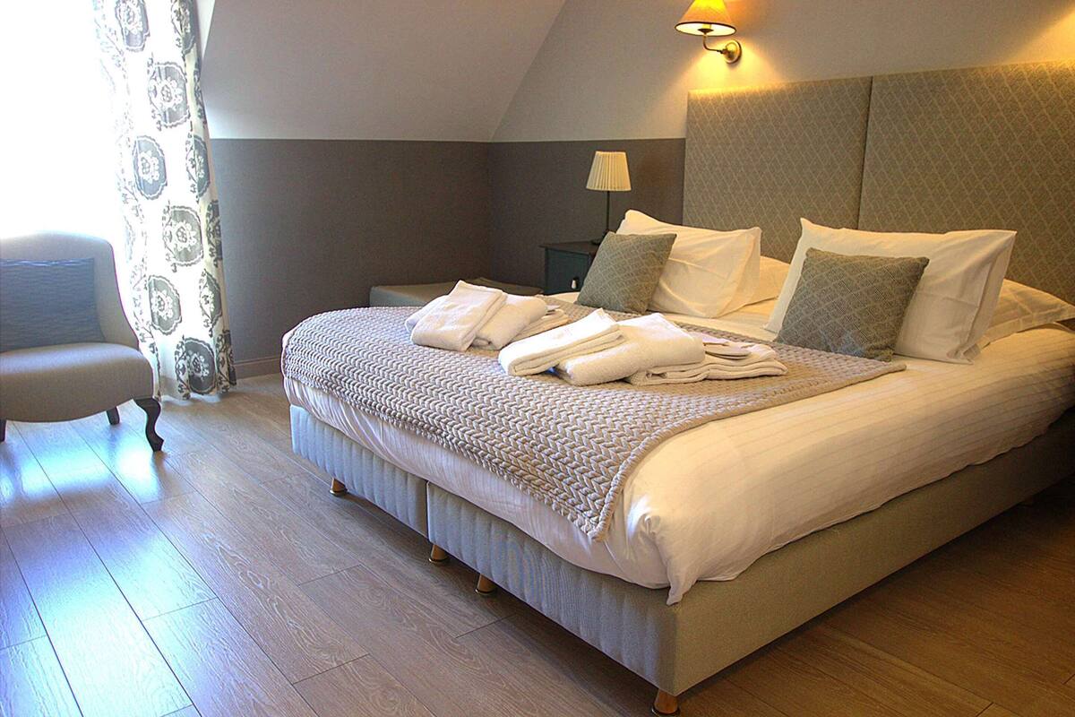 A neatly made bed with pillows and folded towels, a bedside lamp, and a chair in a room with wooden flooring and a patterned curtain.
