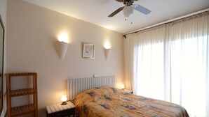 Ein Schlafzimmer mit einem Bett, Nachttischlampen, einem Deckenventilator, einem Holzregal und einem Fenster mit Vorhängen.