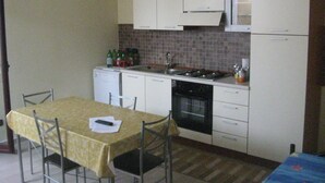 A compact kitchen with white cabinets, a tiled backsplash, a stove, and a sink. There is a dining table with chairs and a small refrigerator.