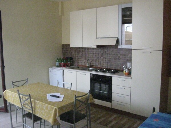 A compact kitchen with white cabinets, a tiled backsplash, a stove, and a sink. There is a dining table with chairs and a small refrigerator.