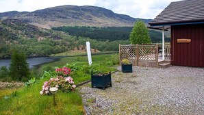 A rustic building named 'Kingfisher' with a gravel path and flower beds, overlooking a river and mountains.