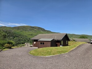 A single-story building with a sloped roof, a gravel driveway, and a grassy area in front. - Kingfisher a beautiful 5 star, 2 bedroom, 2 bathroom holiday property that can sleep up to 4 people (Crianlarich)