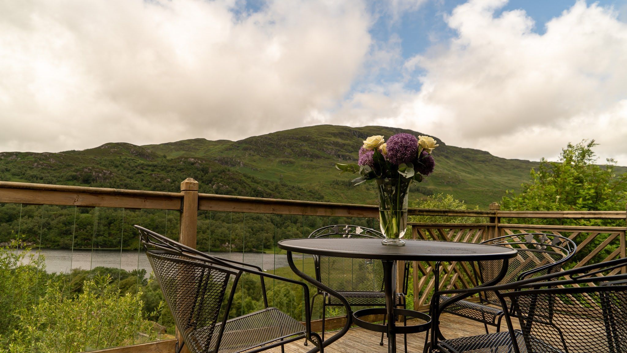 A round metal table with chairs on a deck, overlooking a lake and hills.