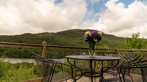 A round metal table with chairs on a deck, overlooking a lake and hills. - Kingfisher a beautiful 5 star, 2 bedroom, 2 bathroom holiday property that can sleep up to 4 people (Crianlarich)