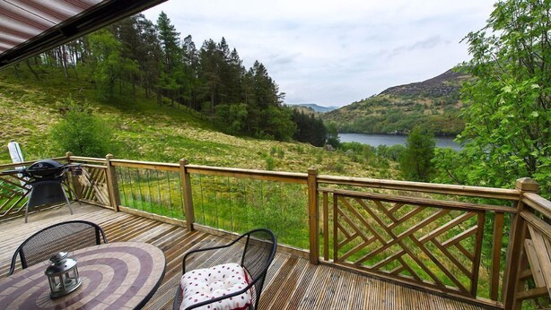 A wooden deck with a table and chairs, overlooking a lake and forested hills.