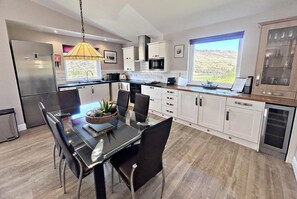 A modern kitchen with a dining area, featuring a stainless steel refrigerator, white cabinetry, and a large window with a view. - Kingfisher a beautiful 5 star, 2 bedroom, 2 bathroom holiday property that can sleep up to 4 people (Crianlarich)