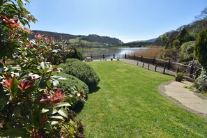 A well-maintained grassy area with a paved walkway, a fence, and a view of a lake with hills in the background. - Charming River Conwy Retreat with Stunning Views (Tal-y-cafn)