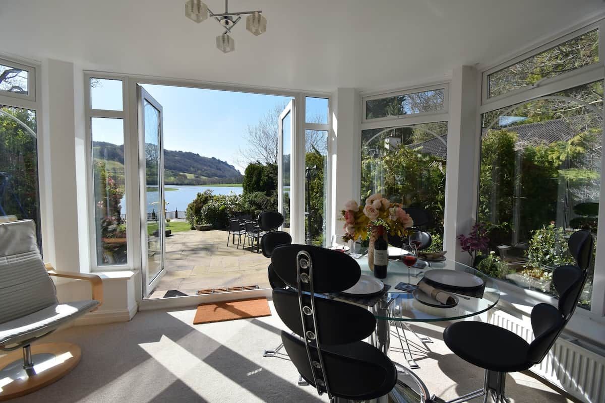 Ein sonnendurchflutetes Zimmer mit einem Glastisch, Stühlen und Blick auf einen See und Hügel.