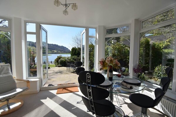 Una habitación soleada con una mesa de vidrio, sillas y vista a un lago y colinas.