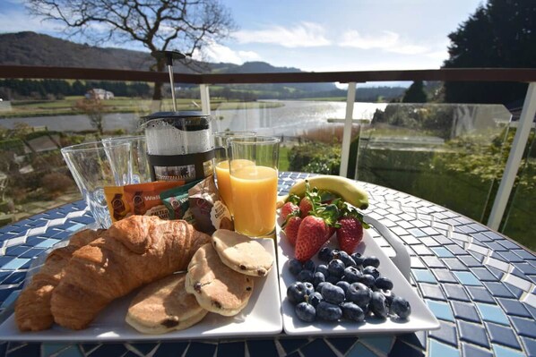 A breakfast table with croissants, pancakes, a plate of strawberries and blueberries, a glass of orange juice, and a coffee maker.