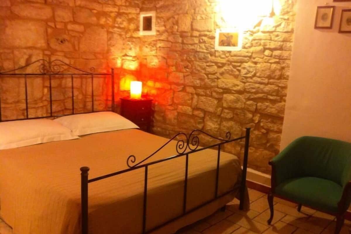 A room with a stone wall, a bed with a metal frame, a nightstand with a lamp, and a green chair.