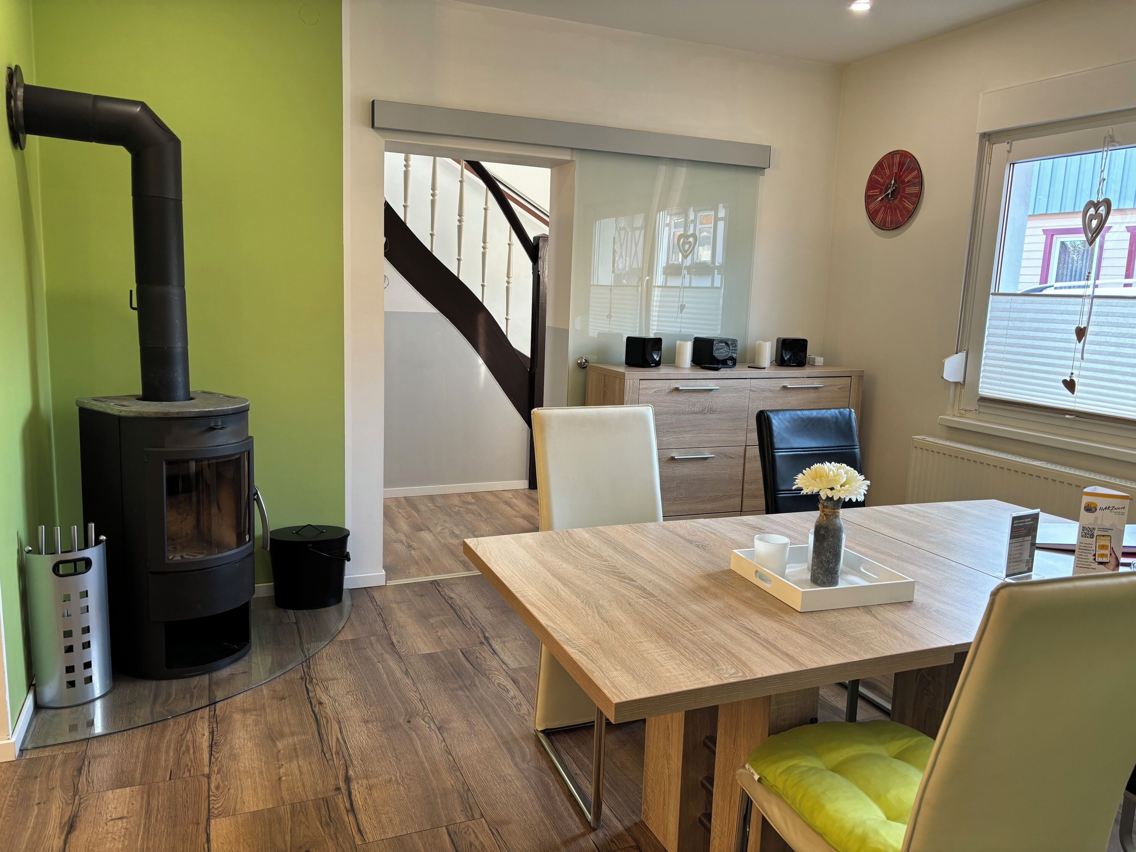 A modern living room with a wood-burning stove, a dining table with chairs, a staircase, and a view of the outside through a window.