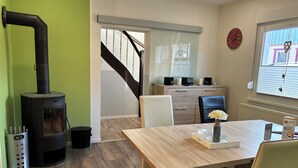 A modern living room with a wood-burning stove, a dining table with chairs, a staircase, and a view of the outside through a window.