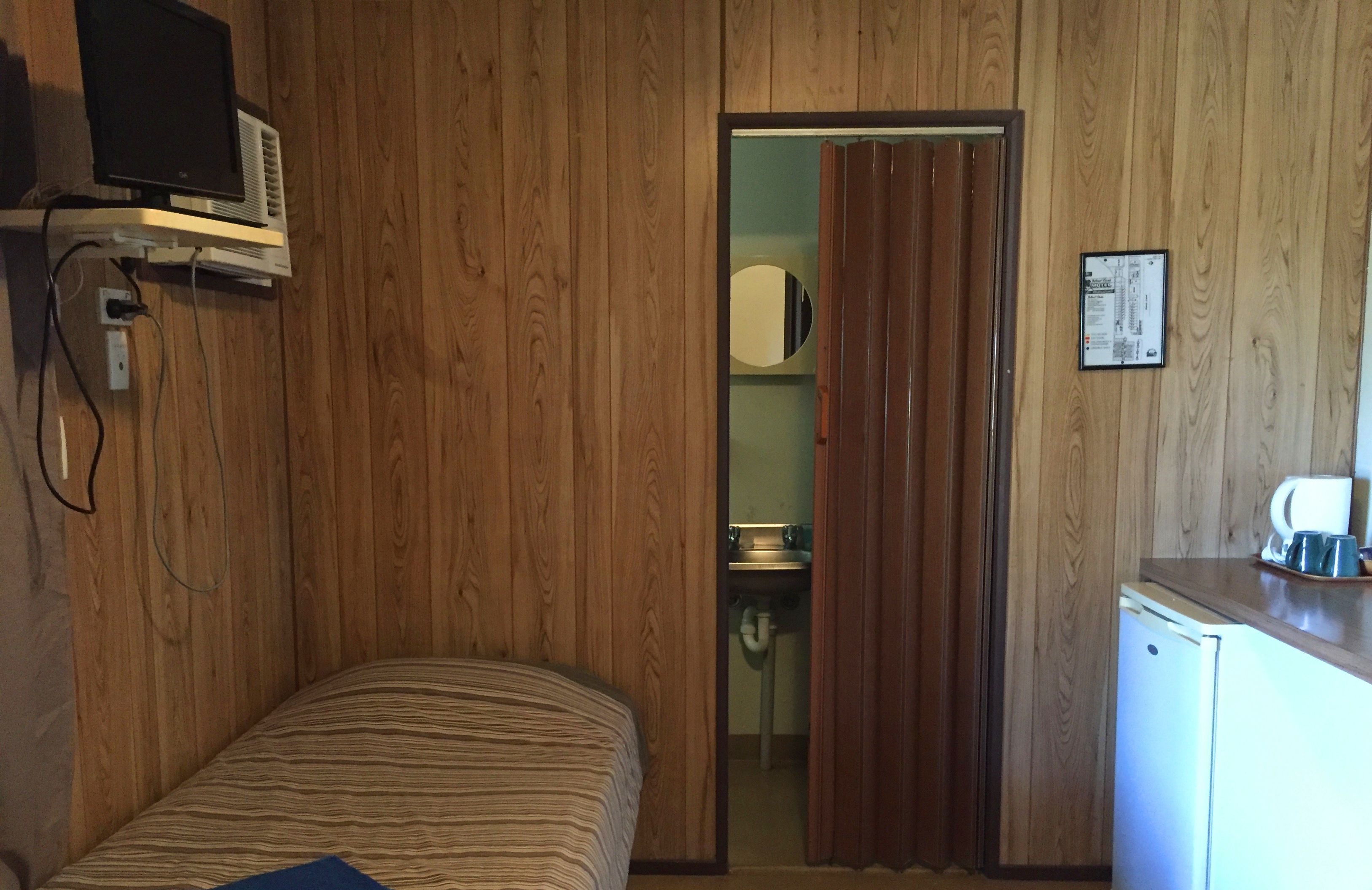 A room with wooden panel walls, a bed with striped bedding, a small kitchenette with a refrigerator, and a television mounted on the wall.