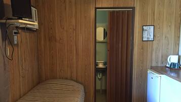 A room with wooden panel walls, a bed with striped bedding, a small kitchenette with a refrigerator, and a television mounted on the wall.