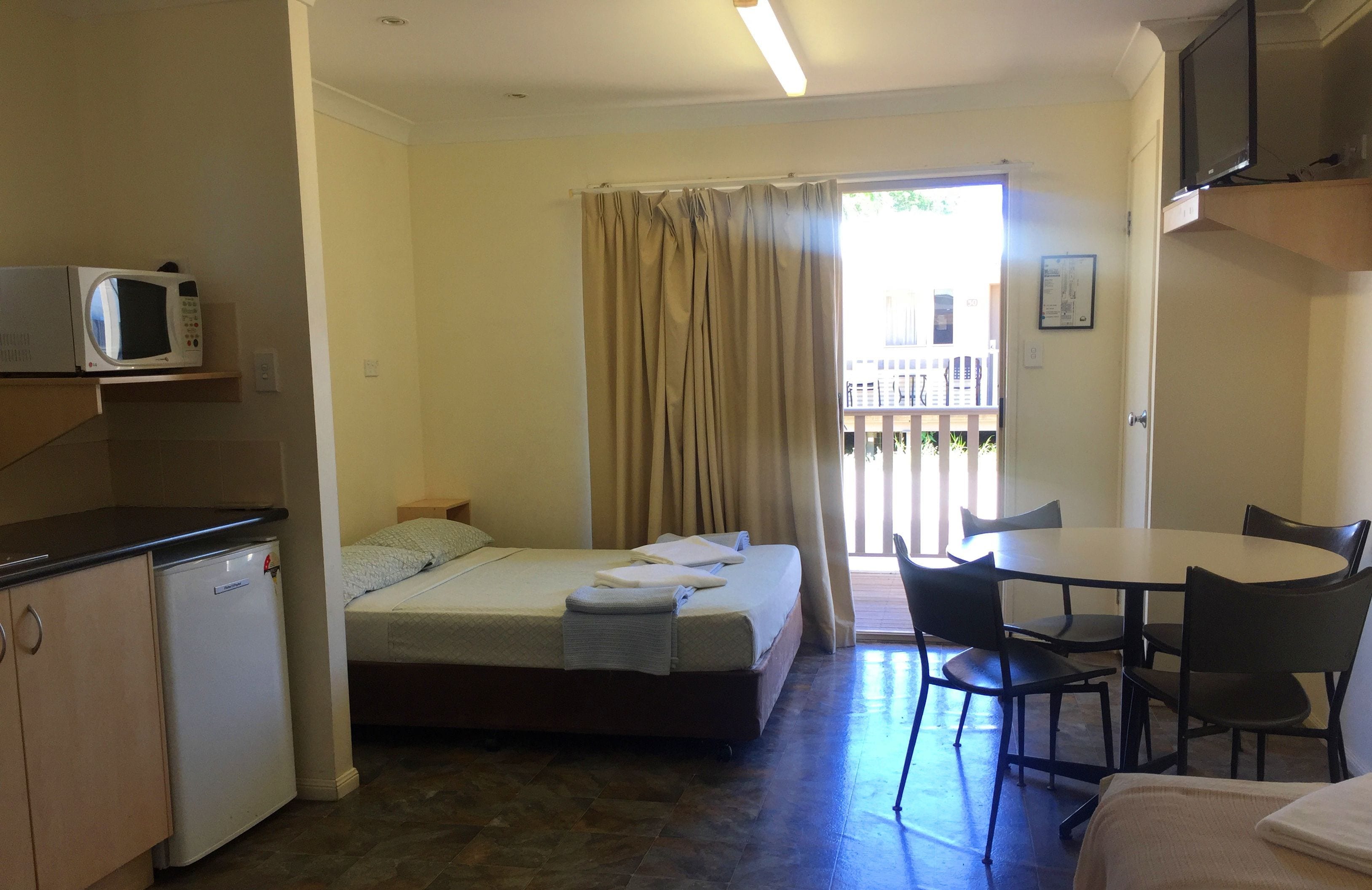 A compact hotel room with a bed, a small kitchenette, a dining table with chairs, and a balcony with a view.