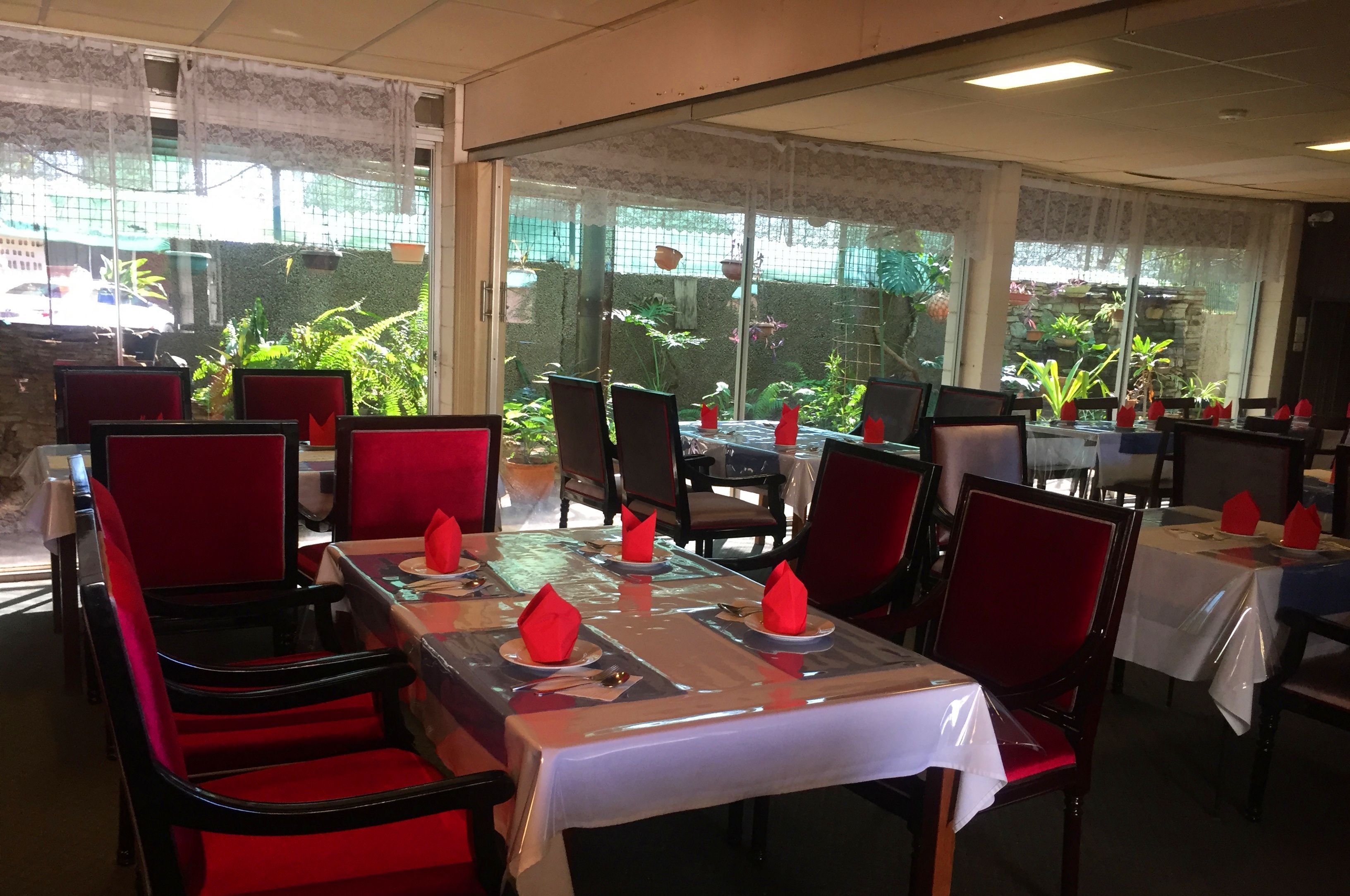 A restaurant with tables set for dining, red chairs, and a view of greenery through the windows.