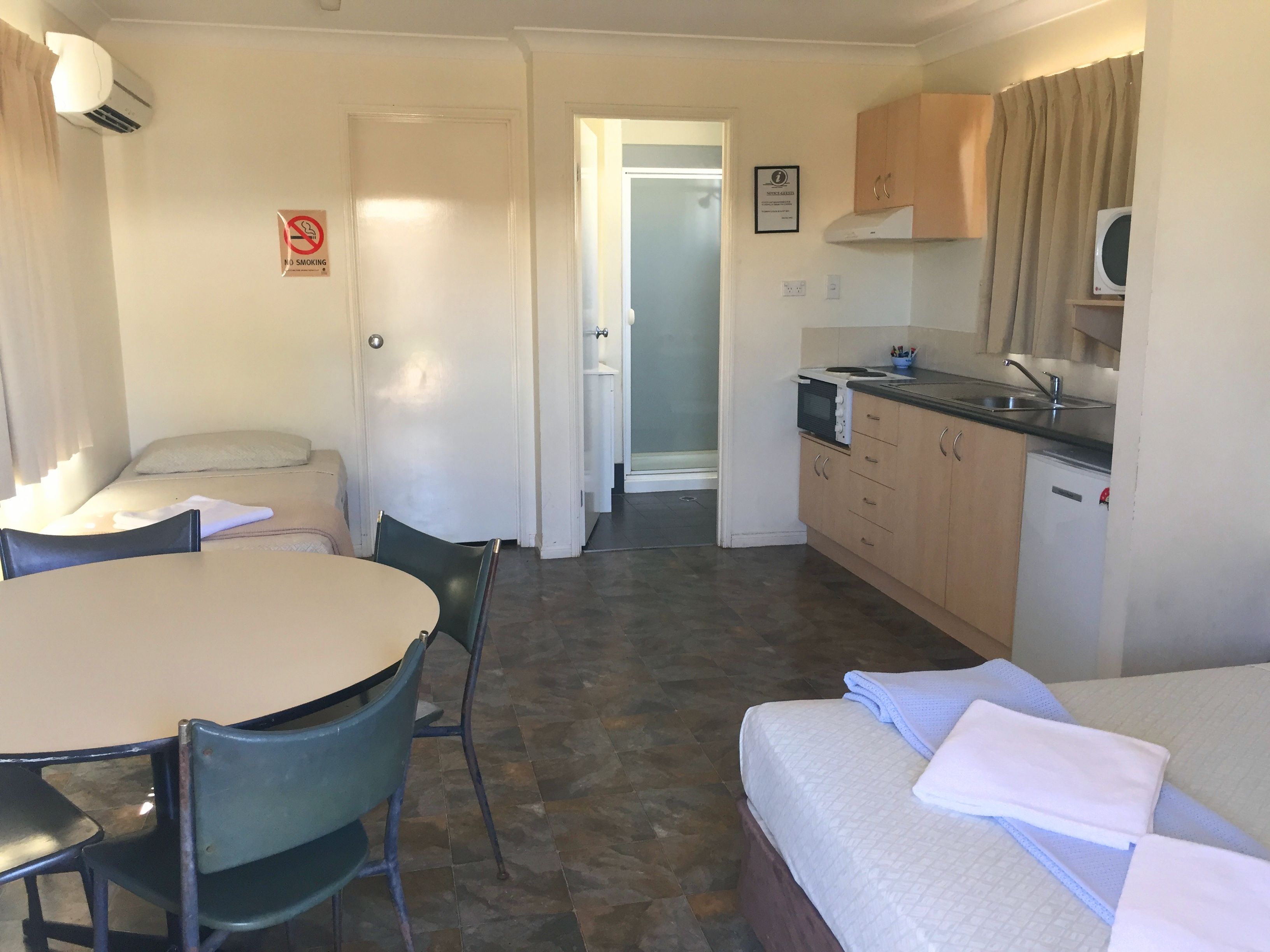 A compact hotel room with a dining table, chairs, a bed with towels, a kitchenette, and a no-smoking sign.