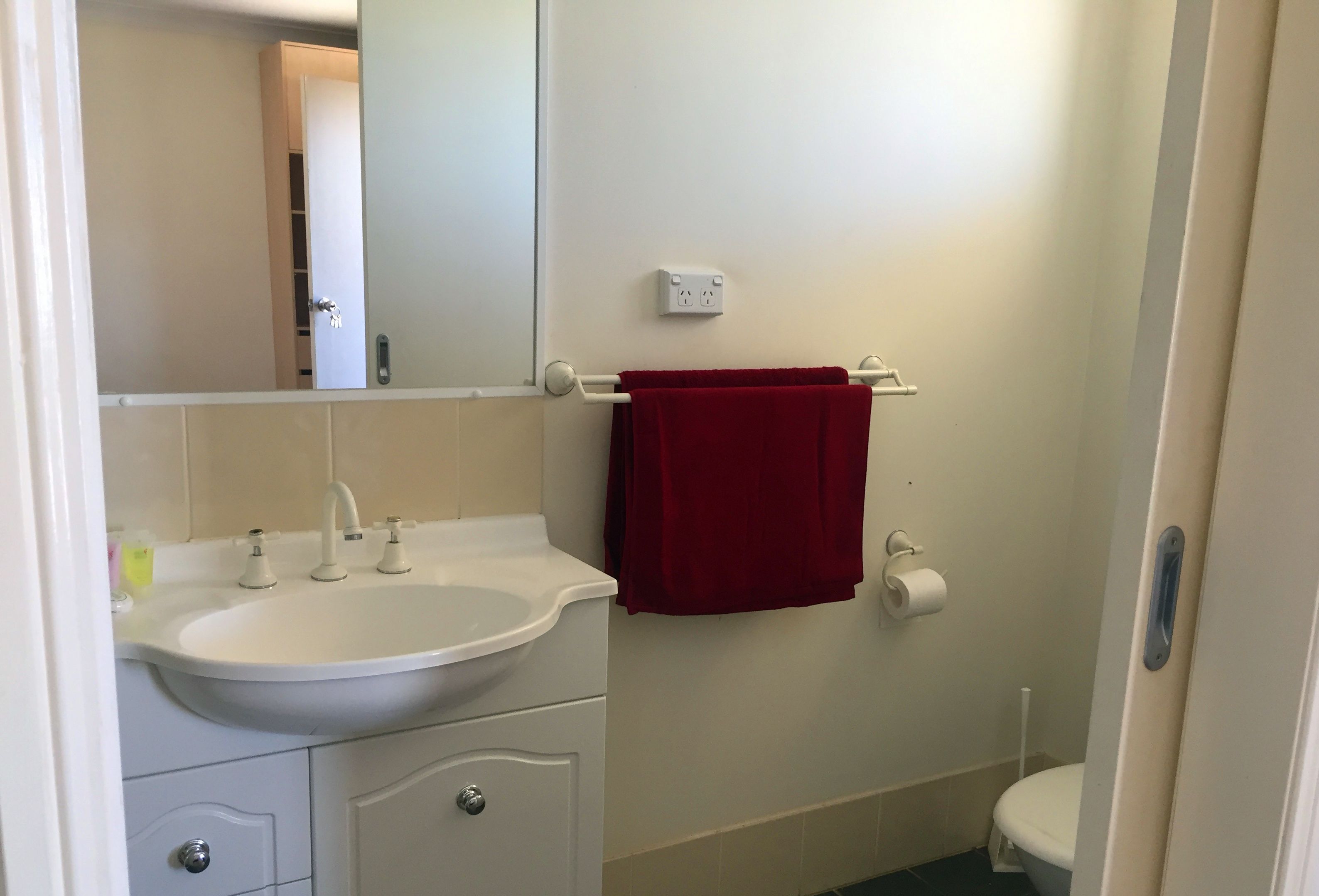 Un baño con un lavabo blanco, una toalla roja colgada en un perchero, un inodoro y un espejo que refleja la puerta.