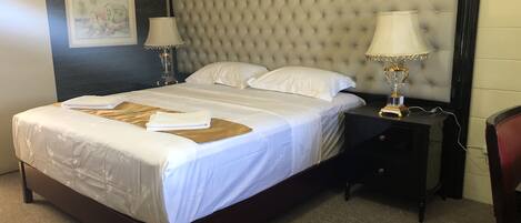 A neatly made bed with white linens and a brown throw, a tufted headboard, two bedside lamps, and a small nightstand with a lamp.